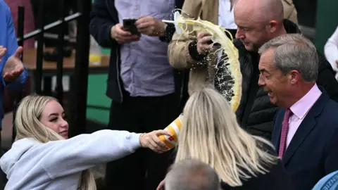 Getty Images A woman in a grey hoody is holding a paper cup in her outstretched hand. There is yellow milkshake coming out of it, flying through the air in the direction of Nigel Farage. He is wearing a dark suit with pink shirt and tie. 